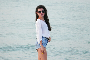 Slim girl walking by the sea. Summer lifestyle image of happy woman walking on the beach. Pretty young woman with long hair  in a white shirt and denim shorts.