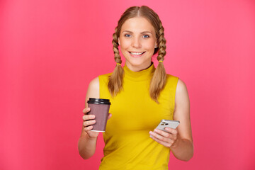 Portrait of beautiful and smiling young woman with pigtails over isolated pink background holding takeaway coffee cup and mobile phone.