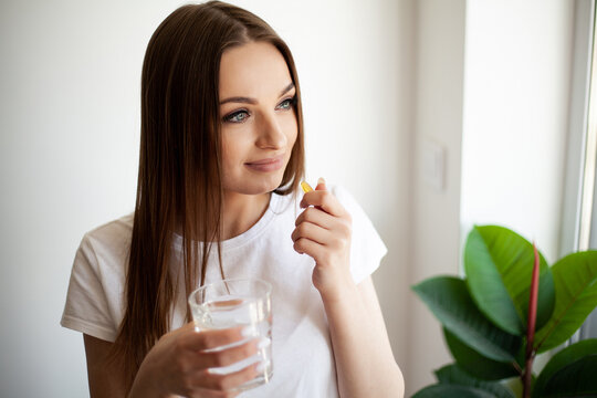 Woman taking vitamin pill with glass of fresh water indoors.