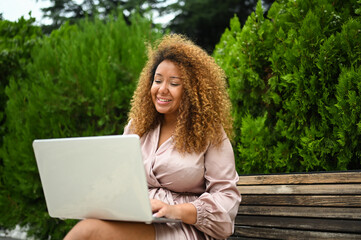 Attractive happy stylish plus size African American woman student freelancer afro hair studying online working on laptop computer outside at summer park. Diversity. Remote work, distance education