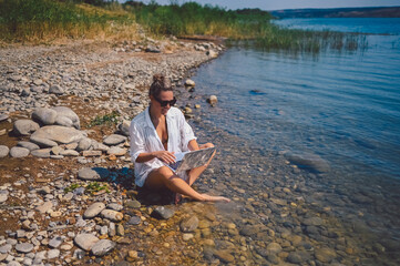 Young woman freelancer traveler wearing white shirt anywhere working online outdoors using laptop enjoying lake view. Happy female downshifter in sunglasses holding computer at sea coastline 