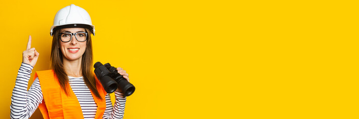 Young woman with a smile in a vest and a helmet holds binoculars on a yellow background....