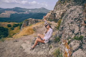 Naklejka premium Young woman freelancer traveler working online anywhere outdoors using laptop enjoying mountain peak view. Happy female downshifter in sunglasses holding computer in unusual workplace at summer