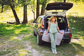 Happy senior woman in straw hat sitting in car trunk or boot outdoors in camper sunny summer park, smiling enjoying journey. Retired people activity and road trip concept. 
