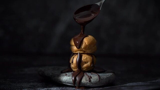 Delicious cream puff sprinkled with melted chocolate. Bakery pastry chef topping liquid chocolate sauce on fresh cream puff or profiterole. Moody background with stone plate.