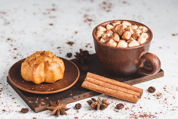 Hot chocolate drink with marshmallows in a cup and sweet bakery eclair dessert sprinkled with cocoa powder, cinnamon stick and star anise on white table