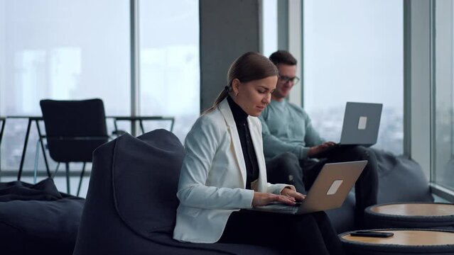 Positive attractive Caucasian lady in white jacket works on laptop she&rsquo;s holding on her laps. Woman smiles, looks at her watch and continues typing. Man sitting at backdrop uses his gadget.
