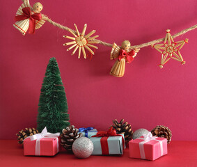 Christmas straw decorations in the shape of a Christmas flake, an angel and a star hanging on a string, Christmas decorations and gifts, pine cones and a green tree on a pink background