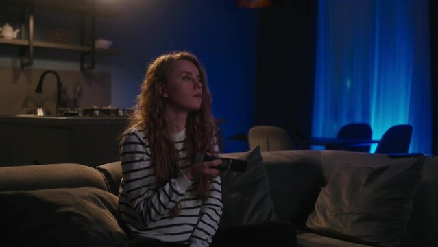 Cute blonde curly woman switching on television, navigating in smart tv applications while choosing channels to watch for entertain herself while sitting on sofa in living room at home. Cinematic shot