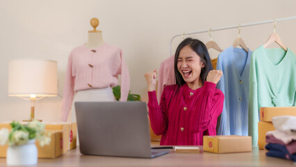 Obraz premium Asian small business owner who is successful in selling online hits the target purchases happy selling on laptop computer. Successful Asian businesswoman raising her hands with joy.