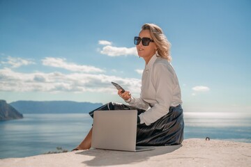 Freelance women sea. She is working on the computer. Good looking middle aged woman typing on a laptop keyboard outdoors with a beautiful sea view. The concept of remote work.
