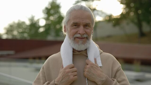 Portrait Smiling Senior Man In Sporty Clothes Looking At Camera Carrying Towel In His Neck Standing In The Park. Elderly Male After Training. Aging Youthfully. People And Sport Concept.