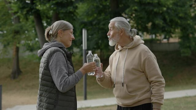 Mature happy family drinking water, prepare for doing sport standing in the park. Elderly couple feeling thirsty. Concept of the couple of pensioners.