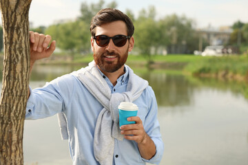 Portrait of handsome bearded man in sunglasses with coffee near lake, space for text