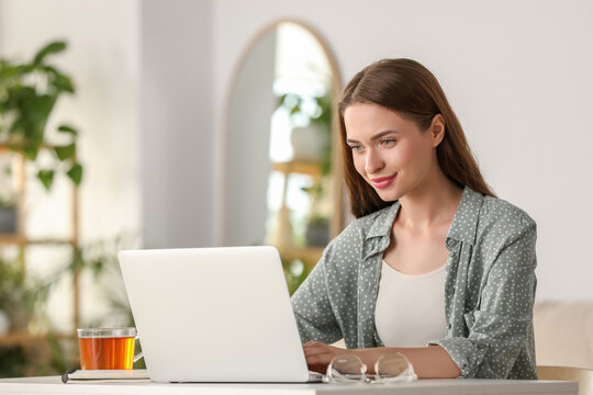 Happy young woman with laptop and tea at table indoors - Powered by Adobe