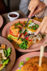 Vertical unrecognizable woman hands use knife, cut tasty, juicy sandwich pizza with omelette fried eggs on plate in cafe