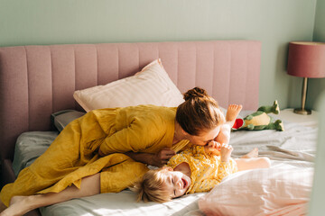 Mom tickles and kisses her little laughing daughter in the morning on the bed in the apartment. - obrazy, fototapety, plakaty