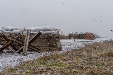 empty roadblock on the road. concrete slabs, sandbags and anti-tank hedgehogs restrict traffic. military structures on a winter road. blizzard and ice on the road at the patrol post. - obrazy, fototapety, plakaty