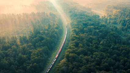 Cargo Train in summer morning forest at fog sunrise. Aerial view of moving freight train in forest....