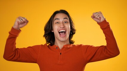 Excited jubilant overjoyed gender fluid man 20s wears orange shirt doing winner gesture celebrate clenching fists say yes isolated on yellow background studio portrait
