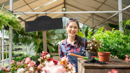 Smiling Florist holding empty wooden sign for texture at garden green house plant shop. Beautiful Asian woman gardener working inside the propagation table using scoop and soil. Signage for words.
