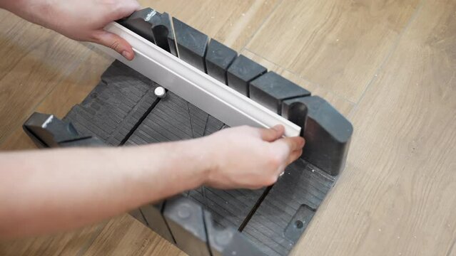 Workman measuring and fixing white plastic plinth for cutting and further installing, handyman using handsaw for cut skirting board on construction site, home baseboard renovation concept
