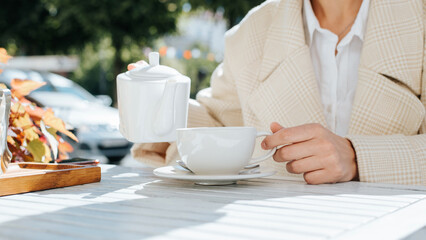 Close-up of woman holding teapot and mug sitting at table in street cafe on sunny day, outdoors. Selective focus on female hand