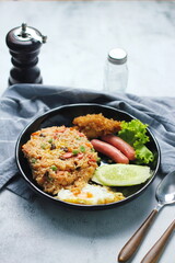 Fried rice with peanuts, carrots, raisins Served with fried chicken, fried egg, and fried sausage. Served on a black plate on a white table in a restaurant.