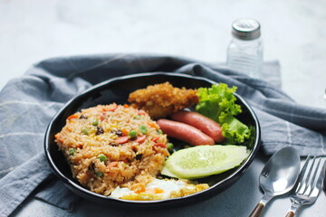 Fried rice with peanuts, carrots, raisins Served with fried chicken, fried egg, and fried sausage. Served on a black plate on a white table in a restaurant.