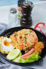 Fried rice with peanuts, carrots, raisins Served with fried chicken, fried egg, and fried sausage. Served on a black plate on a white table in a restaurant.