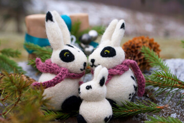 A family of felted rabbits surrounded by branches of a Christmas tree and cones against the backdrop of a New Year's gift and a mountain landscape