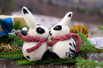 A pair of white and black felted rabbits surrounded by sprigs of a Christmas tree and cones against the background of a New Year's gift and a mountain landscape