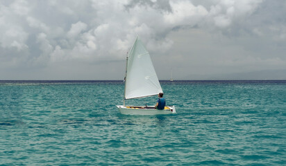 Lonely sailor on training sailing pram optimist education boat in the sea in Greece, water background and cloudy sky - obrazy, fototapety, plakaty