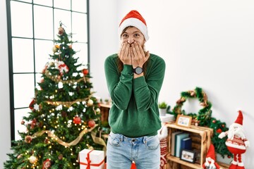 Redhead man with long beard wearing christmas hat by christmas tree laughing and embarrassed giggle covering mouth with hands, gossip and scandal concept