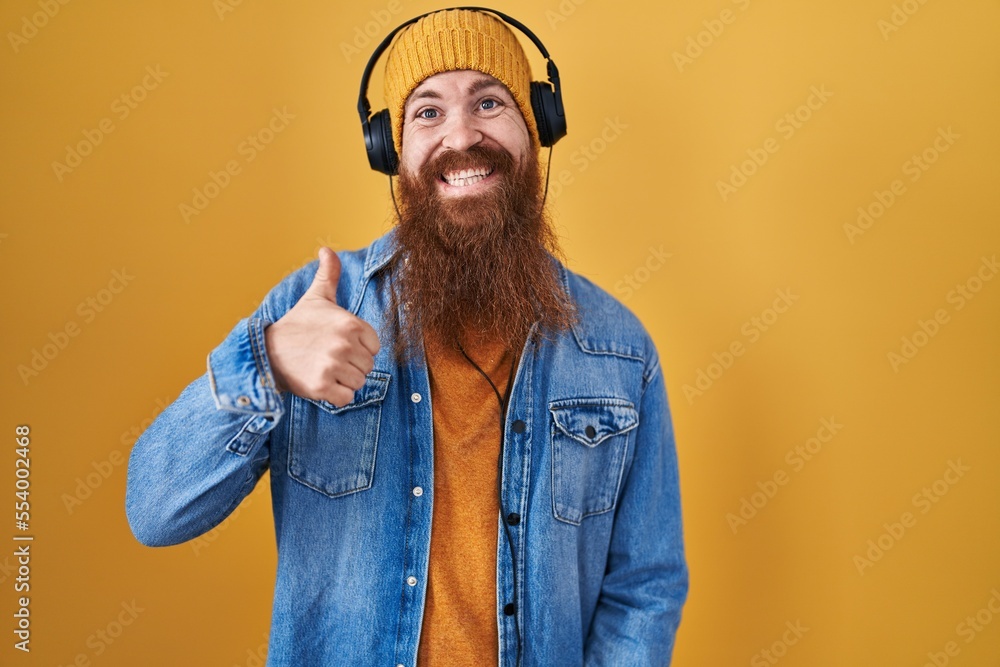 Poster caucasian man with long beard listening to music using headphones doing happy thumbs up gesture with