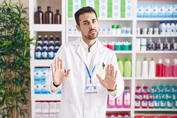Handsome hispanic man working at pharmacy drugstore moving away hands palms showing refusal and...