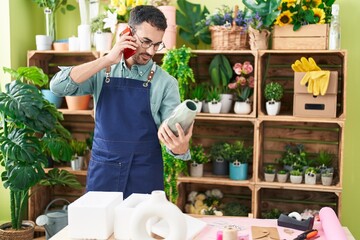 Young hispanic man florist talking on smartphone holding plant pot at flower shop