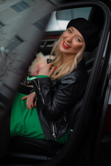 Fototapeta na wymiar Vertical smiling and careless blond woman in black leather jacket, green dress and hat looking at camera from black car