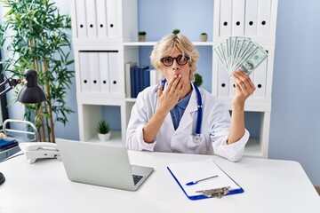 Young doctor man holding money covering mouth with hand, shocked and afraid for mistake. surprised...