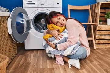 Young caucasian woman hugging clothes sitting on floor at laundry room