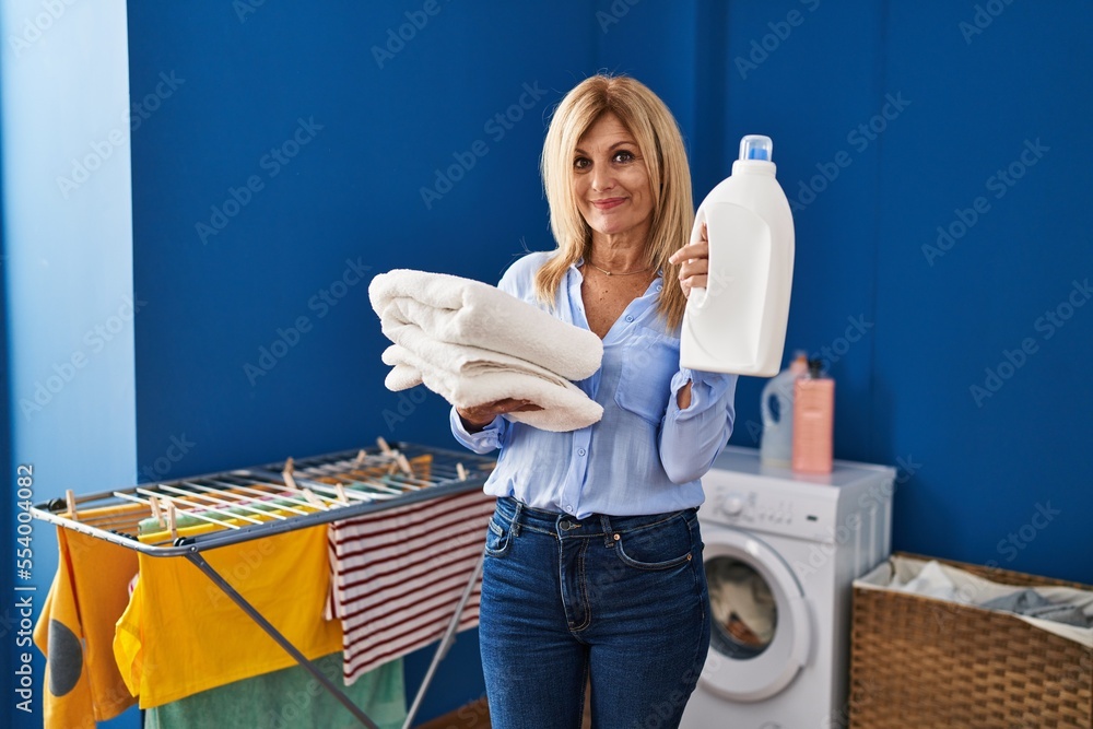 Poster middle age blonde woman holding laundry basket and detergent bottle relaxed with serious expression 