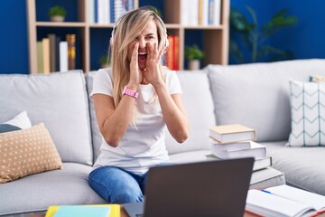 Young blonde woman studying using computer laptop at home shouting angry out loud with hands over mouth