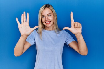 Beautiful blonde woman wearing casual t shirt over blue background showing and pointing up with fingers number seven while smiling confident and happy.