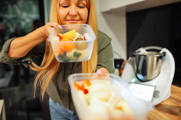 Joyful blond mature woman carrying and smelling fresh vegetables and fruit in plastic containers....