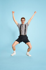 Obraz premium Portrait of young man in checkered shirt, shorts and suspenders showing happiness and excitement isolated over blue backgrounds