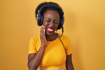 African woman with curly hair standing over yellow background wearing headphones touching mouth with hand with painful expression because of toothache or dental illness on teeth. dentist