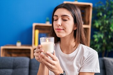 Young beautiful hispanic woman sitting on sofa smelling aromatic candle at home