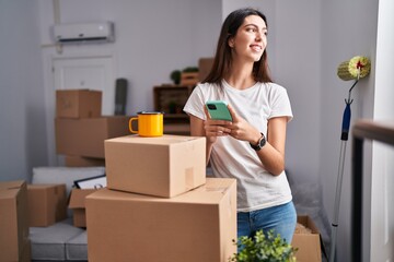 Young beautiful hispanic woman using smartphone drinking coffee at new home