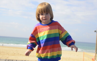 Portrait of a cute boy with long hair. Space for text. The sea and the beach are in the background. A child in a striped multi-colored jacket. Concept: joy, smile, advertising, face close-up.