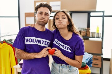 Young hispanic couple wearing volunteer t shirt holding heart puffing cheeks with funny face. mouth inflated with air, catching air.